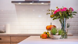 Una encimera de cocina con un florero de flores, un cuenco de fruta y una pequeña planta en maceta.