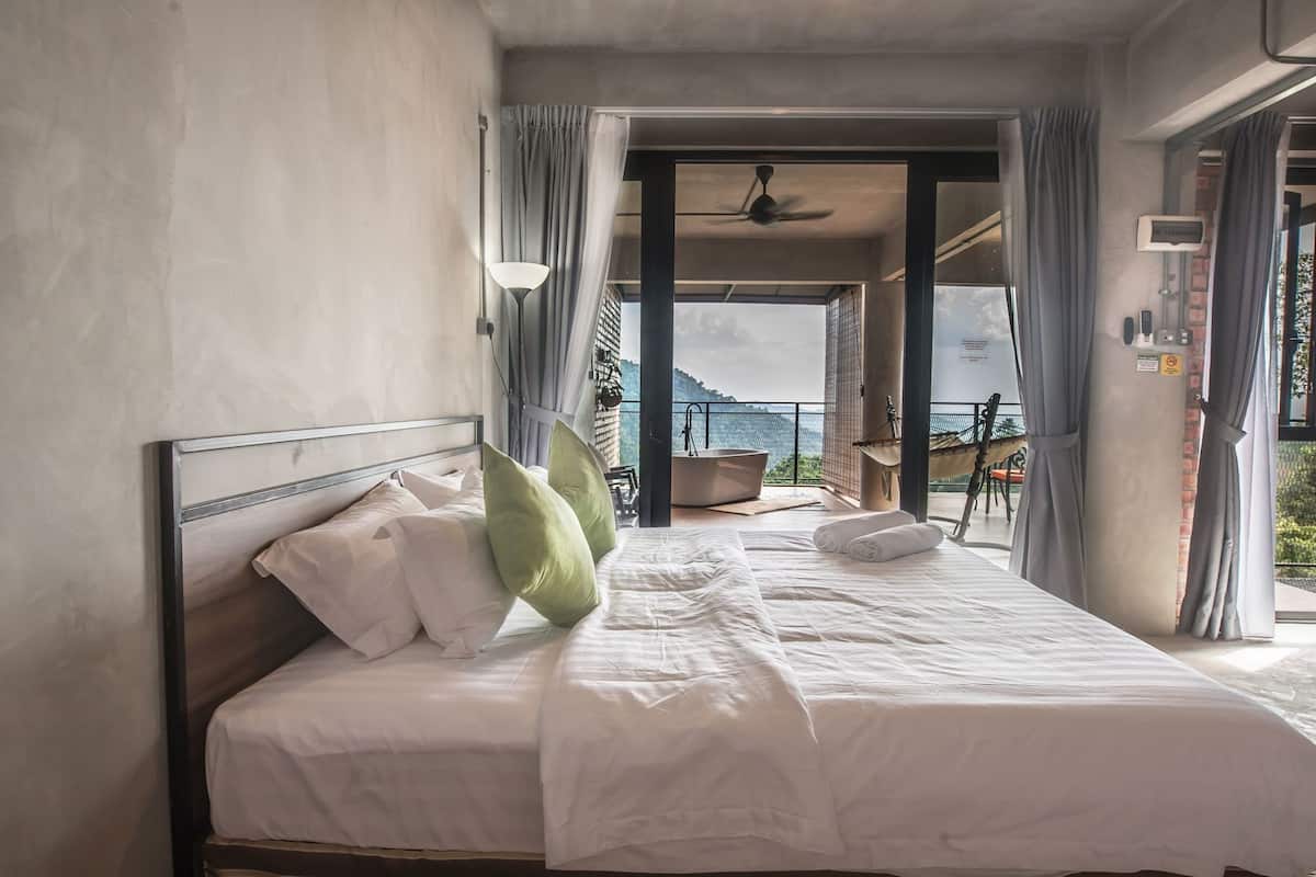 A bedroom with a bed, pillows, a view of the sea, a balcony, and a ceiling fan.