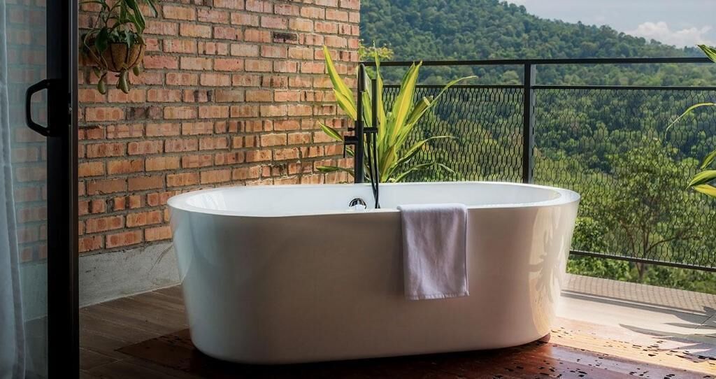 A freestanding bathtub in a room with a brick wall and a balcony overlooking a forested landscape.
