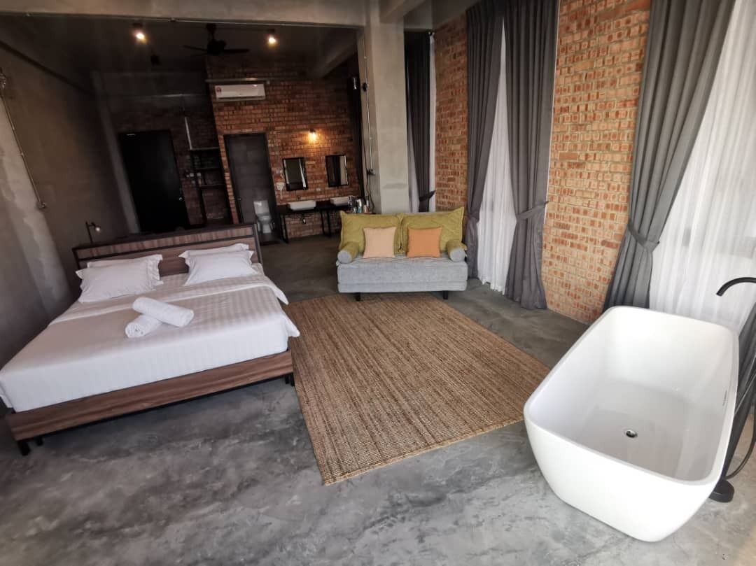 Una habitación de hotel moderna con una cama grande, un rincón de descanso con sofá, una bañera blanca y una zona de cocina al fondo.
