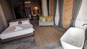 Una habitación de hotel moderna con una cama grande, un rincón de descanso con sofá, una bañera blanca y una zona de cocina al fondo.