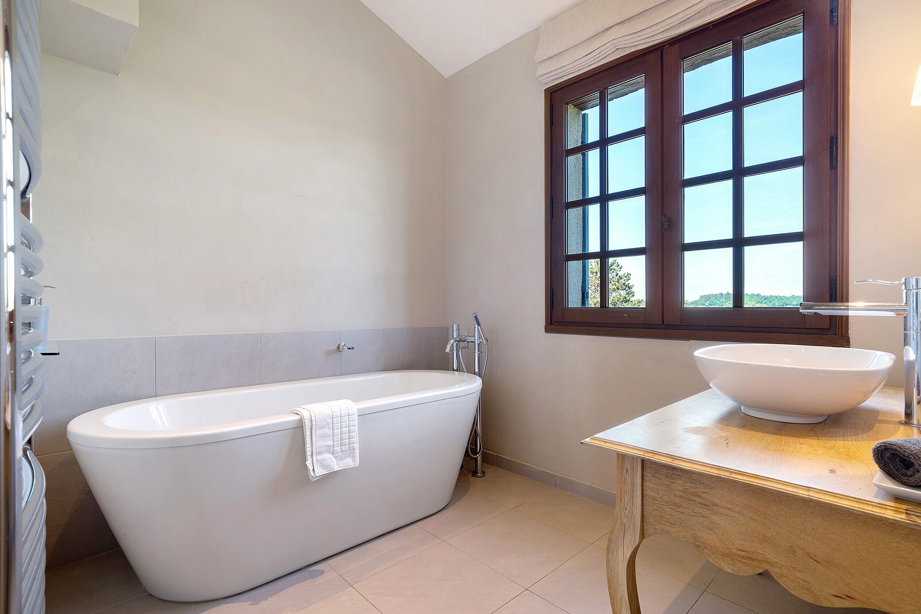 A modern bathroom with a freestanding bathtub, a large window with a view, and a wooden vanity with a bowl sink.