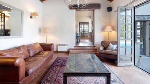 A spacious living area with a leather sofa, a glass-top coffee table, a chandelier, and a view through an open door. - La Maison – Spacious Country Home with Pool and Lush Garden (Villeneuve de Duras)