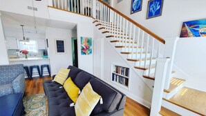 Una sala de estar moderna con un sofá gris oscuro, cojines amarillos y un tapete estampado. Una escalera con barandillas de madera conduce a un nivel superior.
