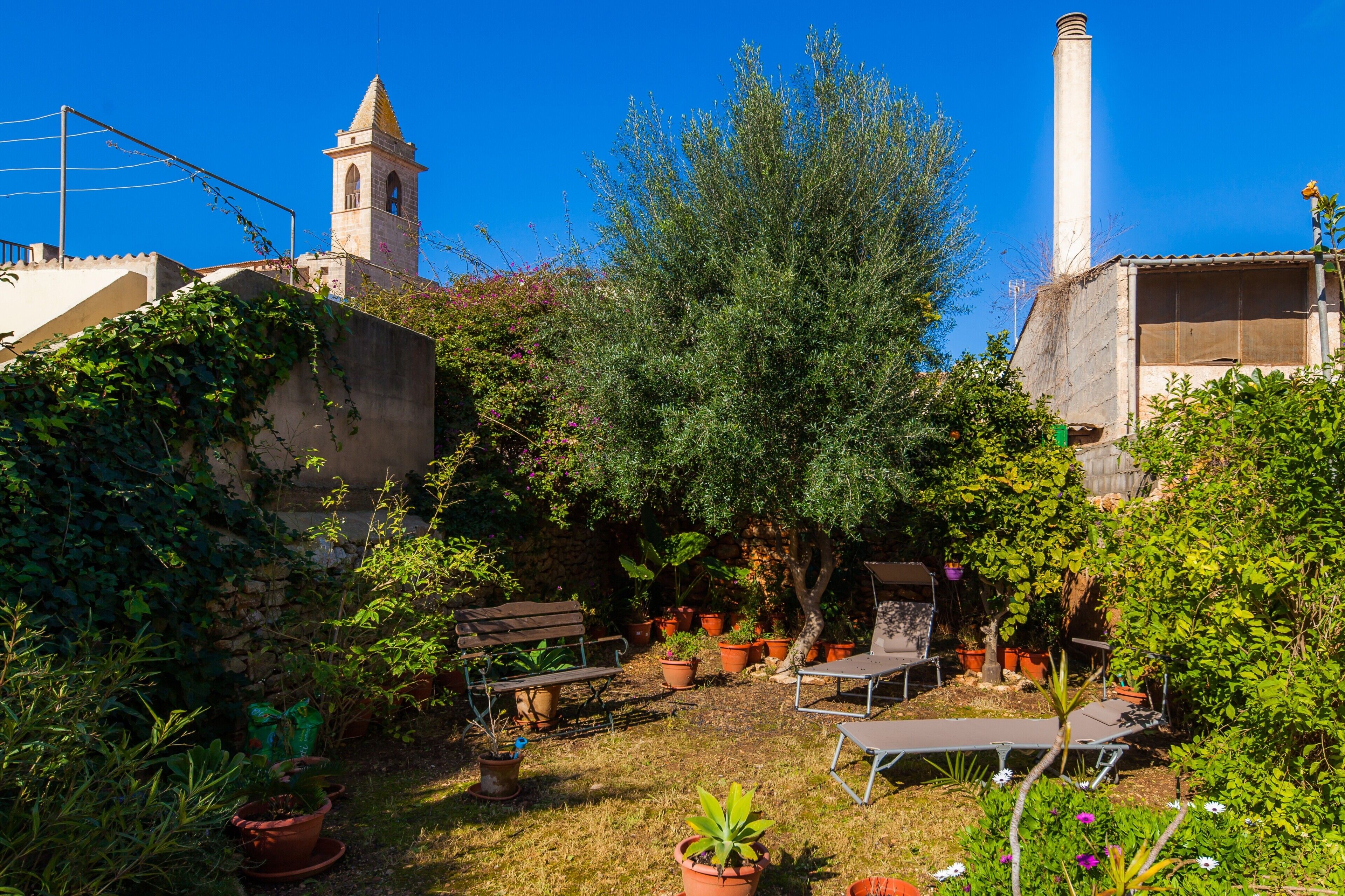 A courtyard with a bench, potted plants, a small table, and a view of a tower and chimney.