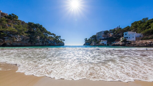 Ein Strand mit klarem Wasser, Gebäuden am Ufer und strahlender Sonne.