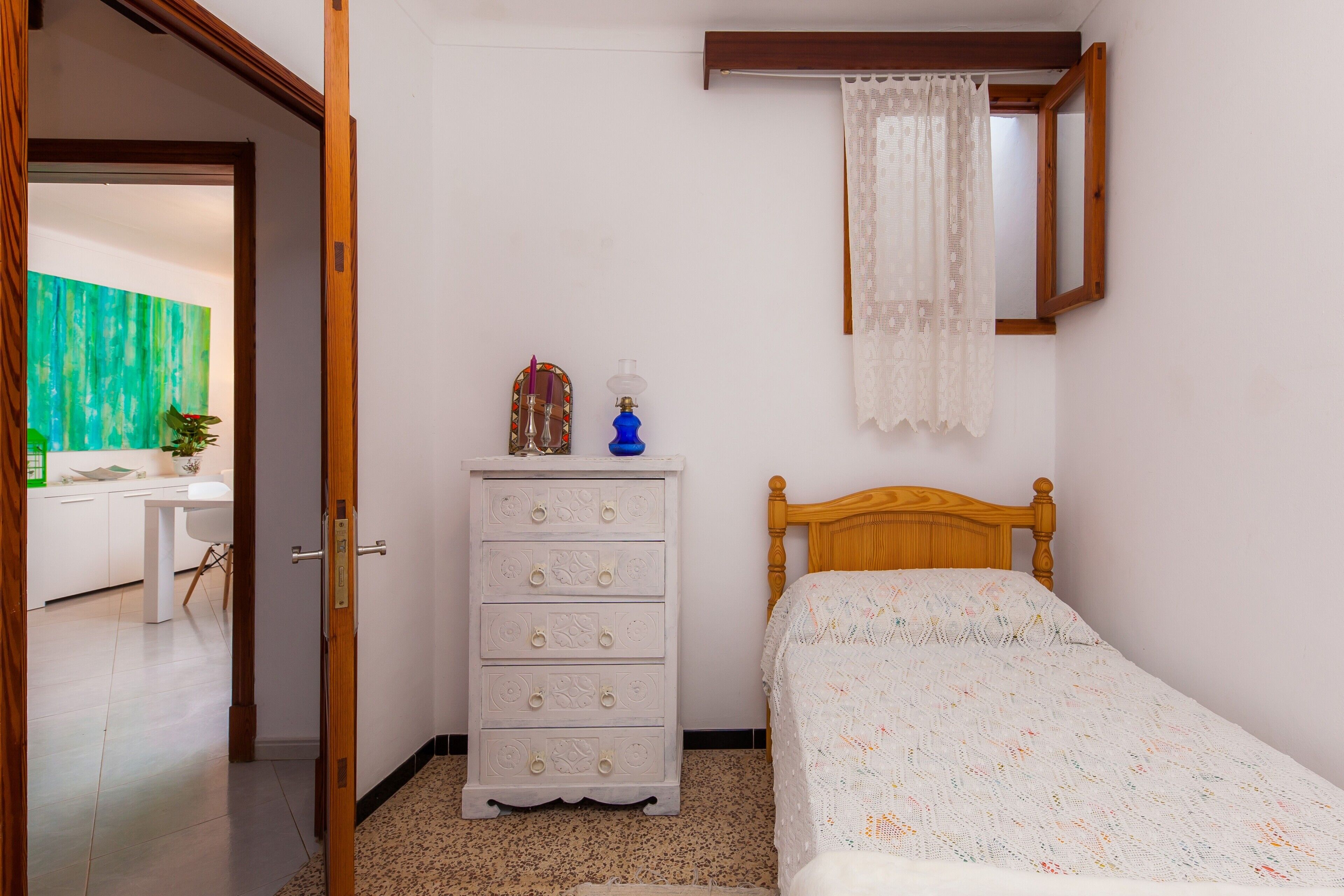 Un dormitorio con una cama de madera, un aparador blanco, un espejo y una ventana con cortina blanca.