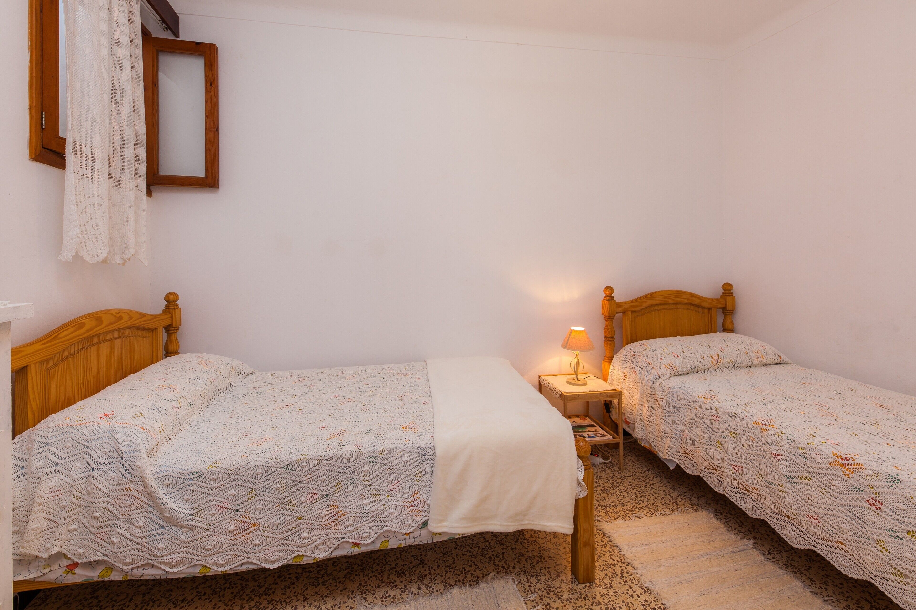 A room with two wooden beds, a nightstand with a lamp, a window with a wooden frame, and a wall-mounted mirror.