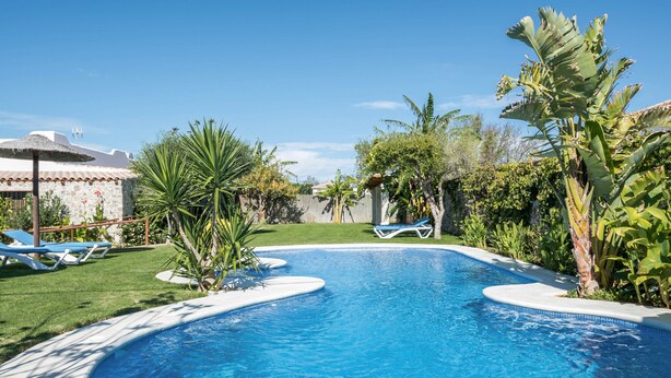 A swimming pool with a curved edge, surrounded by a well-maintained lawn and various tropical plants. - Charming Apartment with Beautiful Pool, Jacuzzi, Patio, & Wi-Fi (CONIL DE LA FRONTERA)