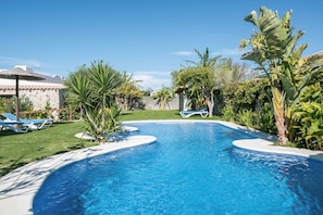 Una piscina de borde curvo, rodeada por un césped cuidado y diversas plantas tropicales.