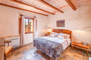 A bedroom with a bed, a nightstand, a wooden bench, a radiator, and a window with curtains. - Secluded Holiday Home Son Garrot with Garden, Mountain View & Wi-Fi; Parking Available (Santanyí)