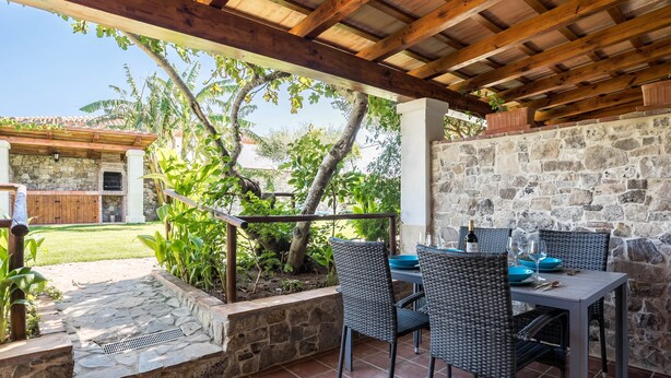 Een terras buiten met stenen muren, een houten pergola en rieten meubilair.