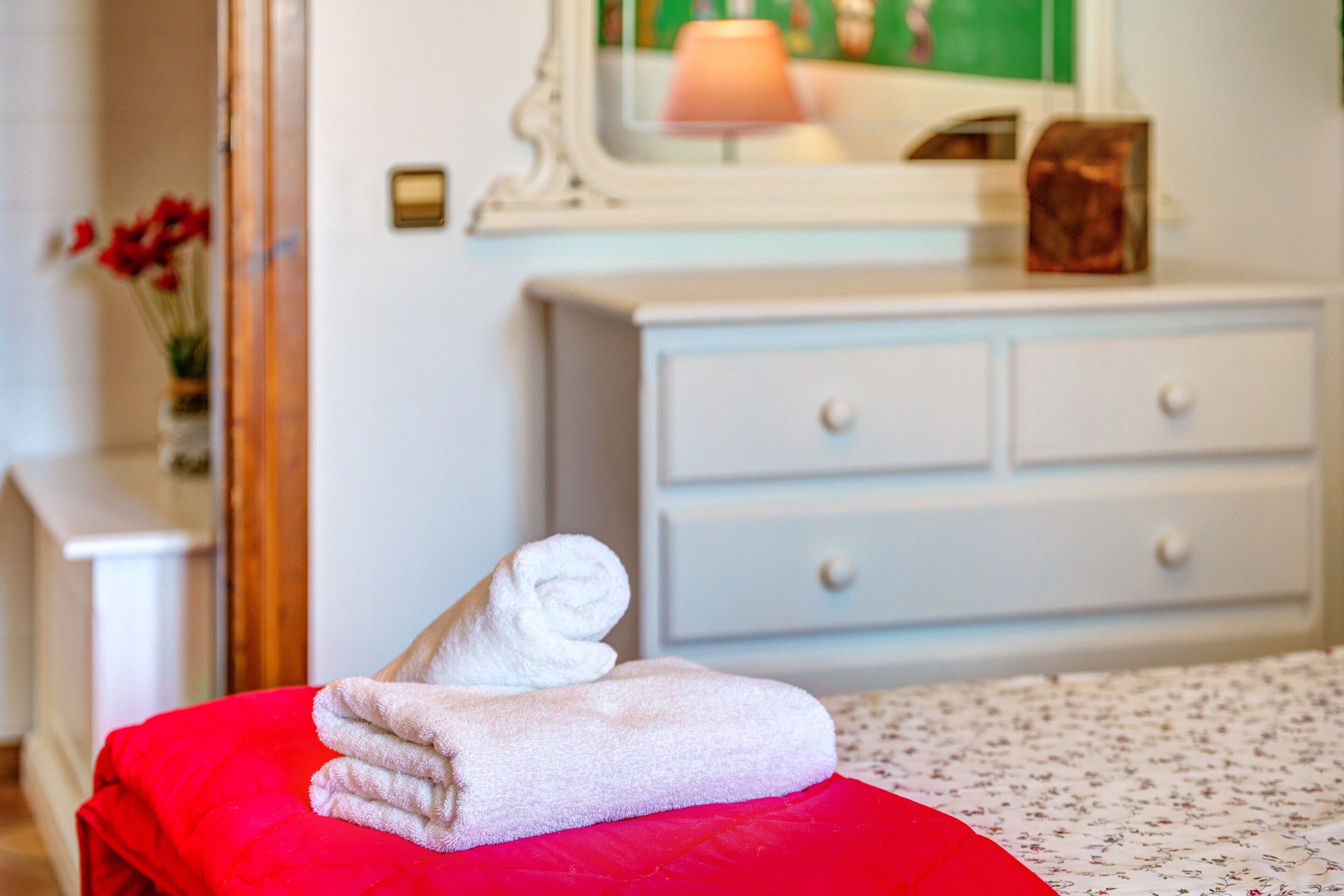 A neatly made bed with a red pillow and a rolled white towel on it.