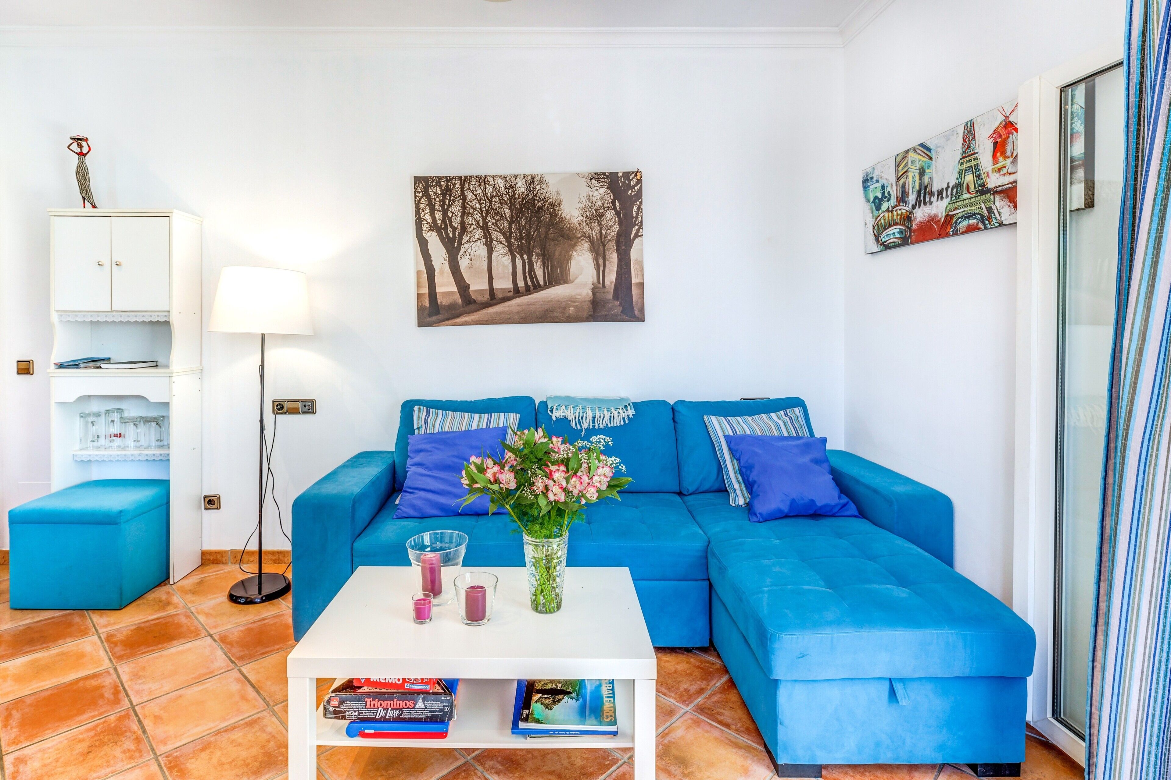 A living room with a blue sofa, a white coffee table with books, a vase with flowers, and a painting on the wall.