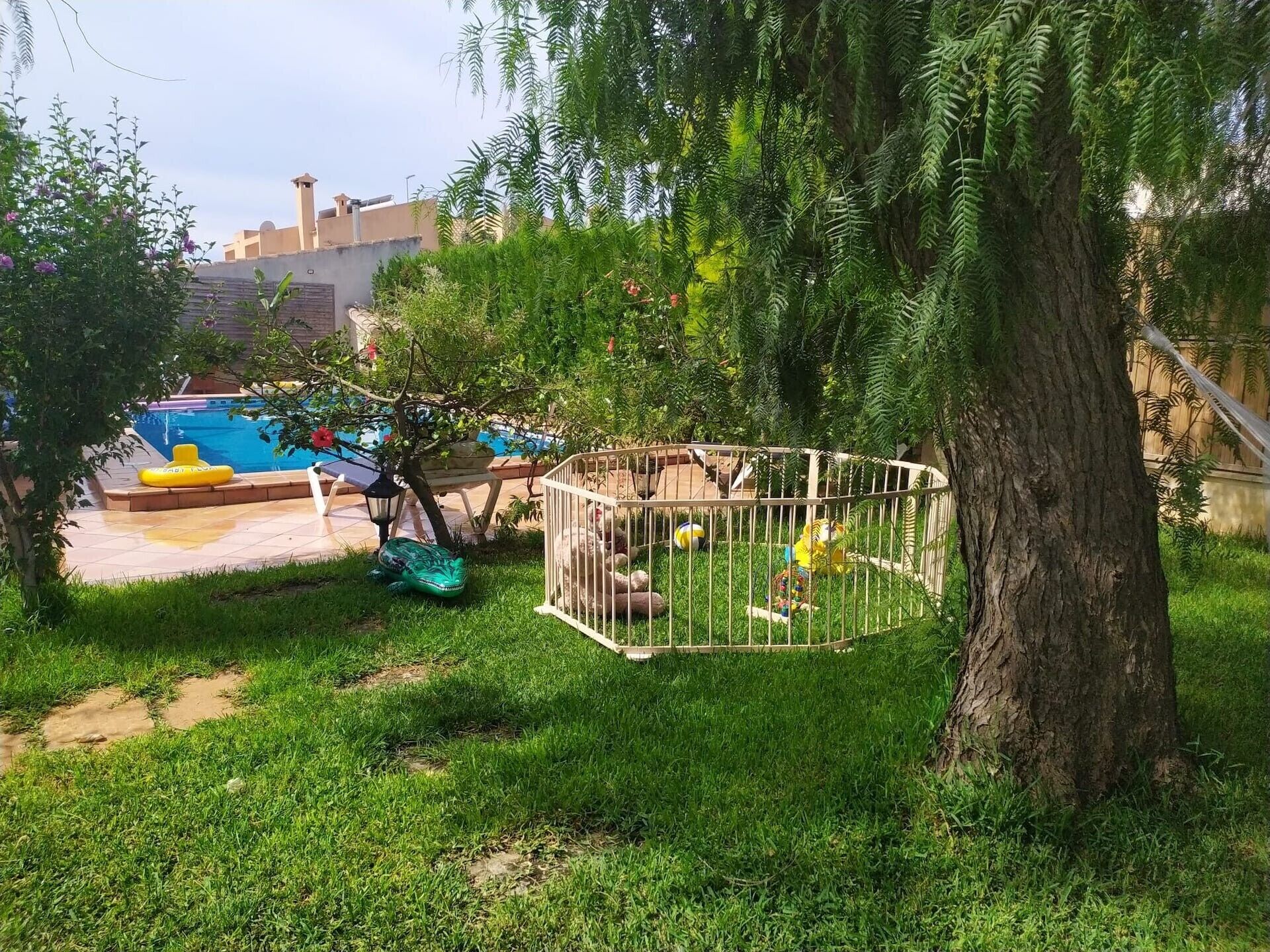 Un patio trasero con piscina, área de juegos cercada y un árbol.