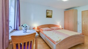 A bedroom with a bed, bedside tables, a desk with a chair, a window with curtains, a wooden wardrobe, and a framed picture on the wall.