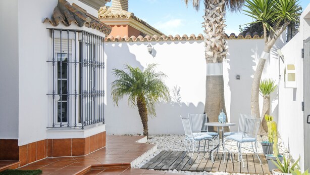 Un cortile con pavimento piastrellato, una parete bianca, un cancello in metallo, una palma e un tavolo con sedie.