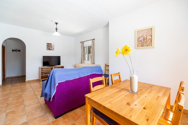 A room with a bed, a TV, a dining table, and a vase with flowers.