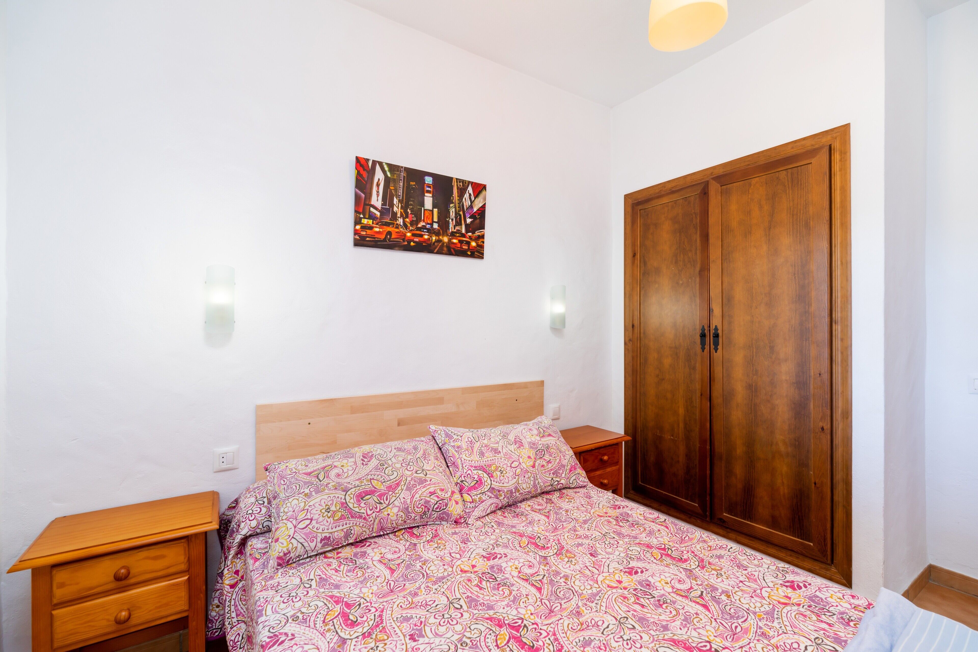 Un dormitorio con cama, armario de madera, mesitas de noche y un cuadro de un paisaje urbano en la pared.