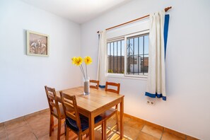 A dining area with a wooden table and chairs, a vase with flowers, a window with curtains, and a wall-mounted picture.