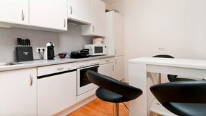 Cocina compacta con gabinetes blancos, microondas, cafetera y estufa. Hay un pequeño comedor con una mesa blanca y dos sillas negras.