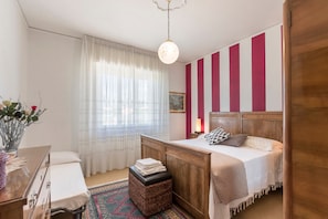 Une chambre avec un lit en bois, une table de chevet, un tabouret en osier, une fenêtre avec des rideaux légers et un mur à rayures verticales.