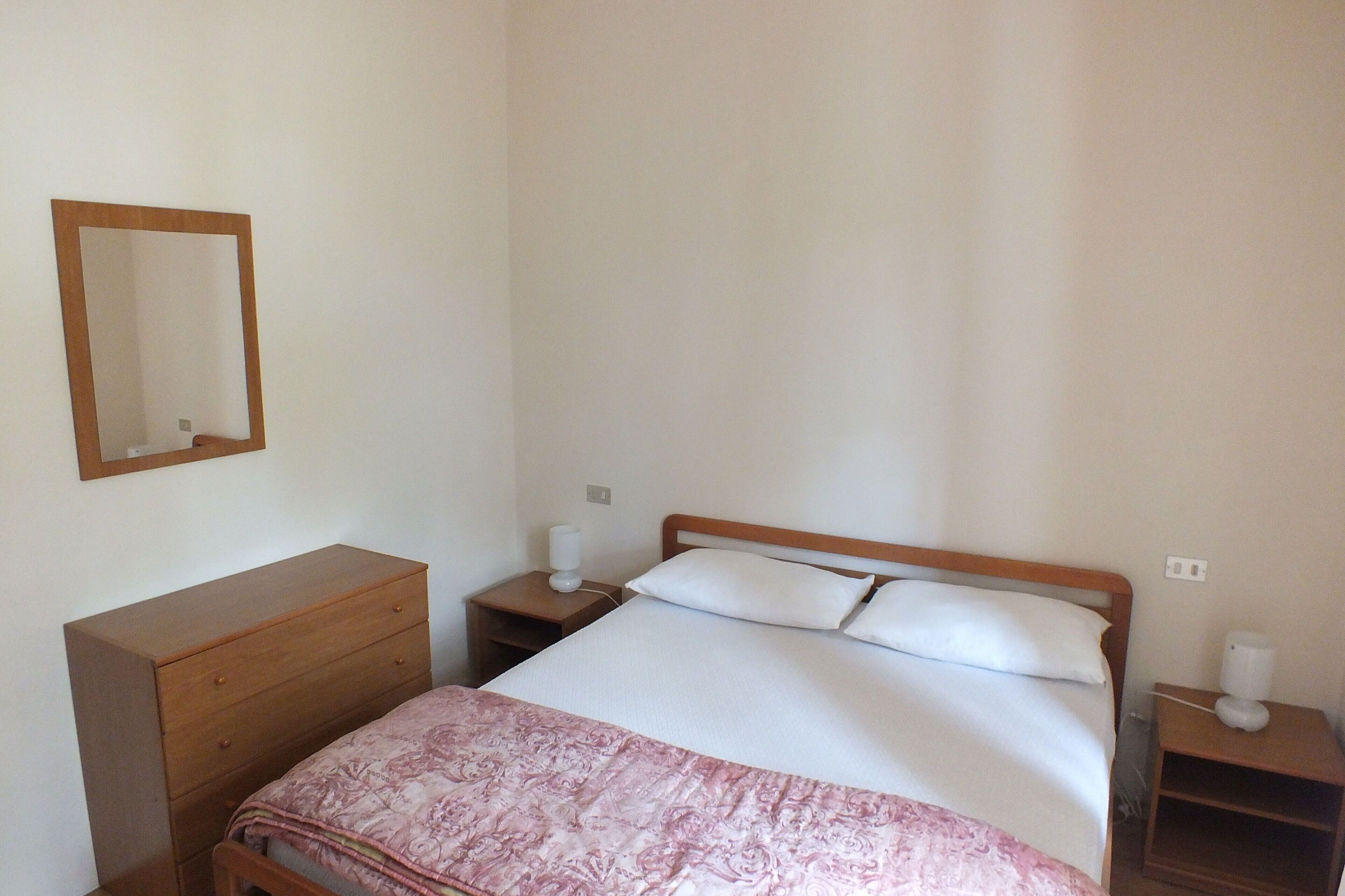 Una camera da letto con un letto, una cassettiera in legno, un comodino con una lampada e uno specchio appeso al muro.