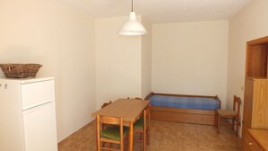 Pieni huone, jossa on puinen pöytä, tuoleja, penkki, pieni jääkaappi ja riippuvalaisin.