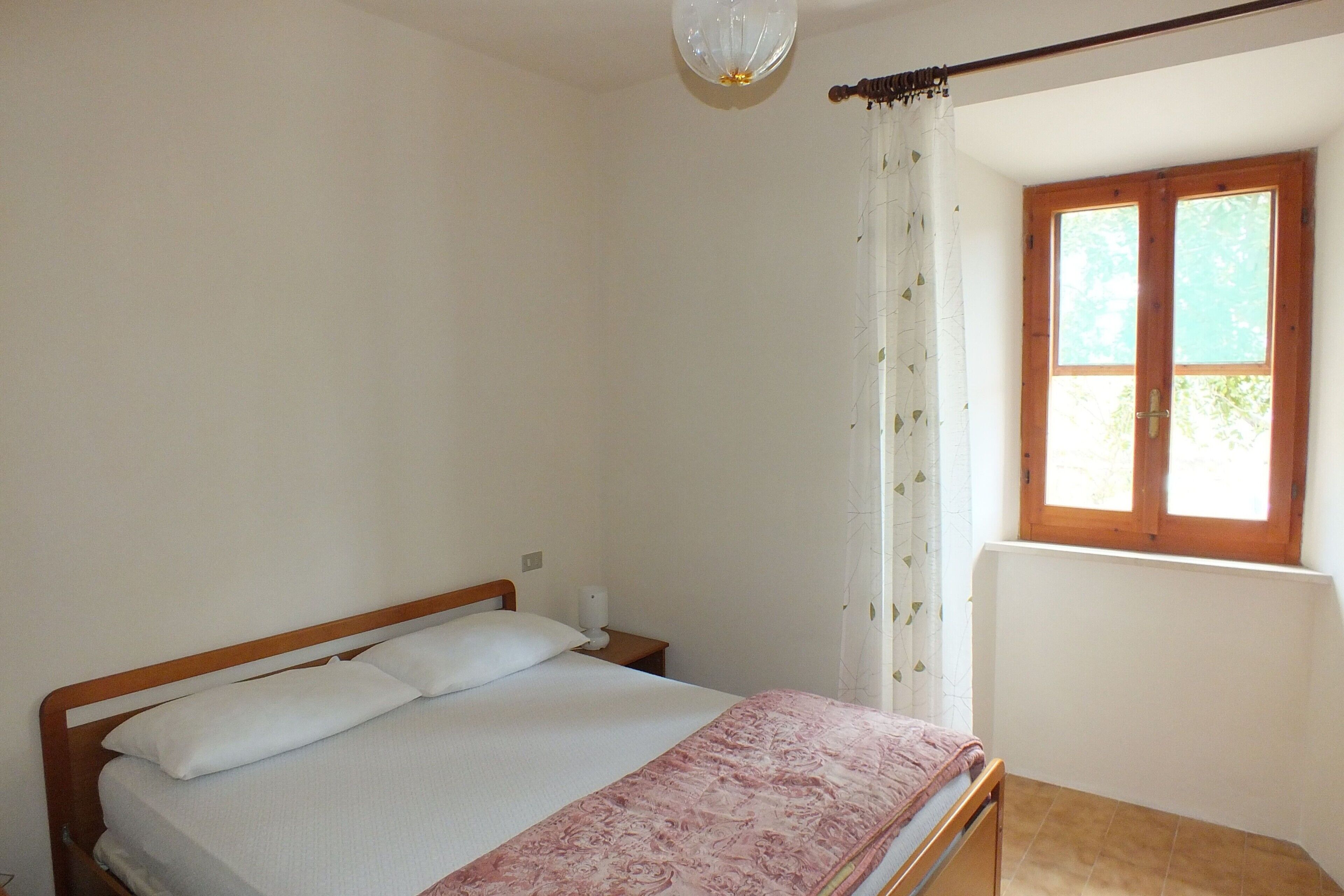 Una camera da letto con un letto, un comodino, una finestra con tende e una lampada a soffitto.
