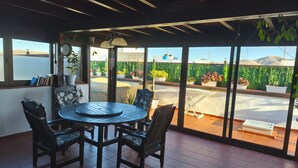 A patio area with a round table and chairs, a grill, and a view of a green hedge and mountains. - Apartment "Casa Orymar" with Ocean View, Mountain View, Balcony & Terrace (El Cotillo)