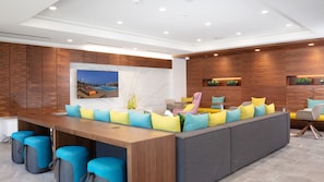 Un moderno vestíbulo de hotel con un mostrador de recepción de madera, asientos coloridos y un televisor de pared grande que muestra una escena de piscina.