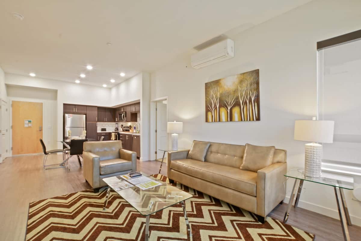 A modern living room with a sofa, coffee table, and a rug. There is a kitchen in the background with cabinets and a refrigerator.