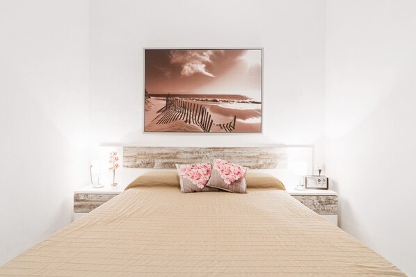 Une chambre à coucher avec un lit, des oreillers, une table de chevet, une commode, un tableau accroché au mur et une fenêtre.