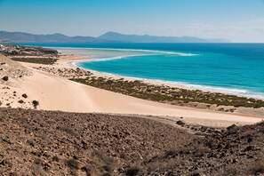 A coastal landscape with a sandy beach, a clear blue sea, and distant mountains.