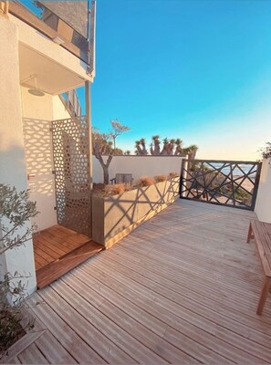 Um deck de madeira com vista para o mar, uma parede branca com treliça e um banco de madeira.