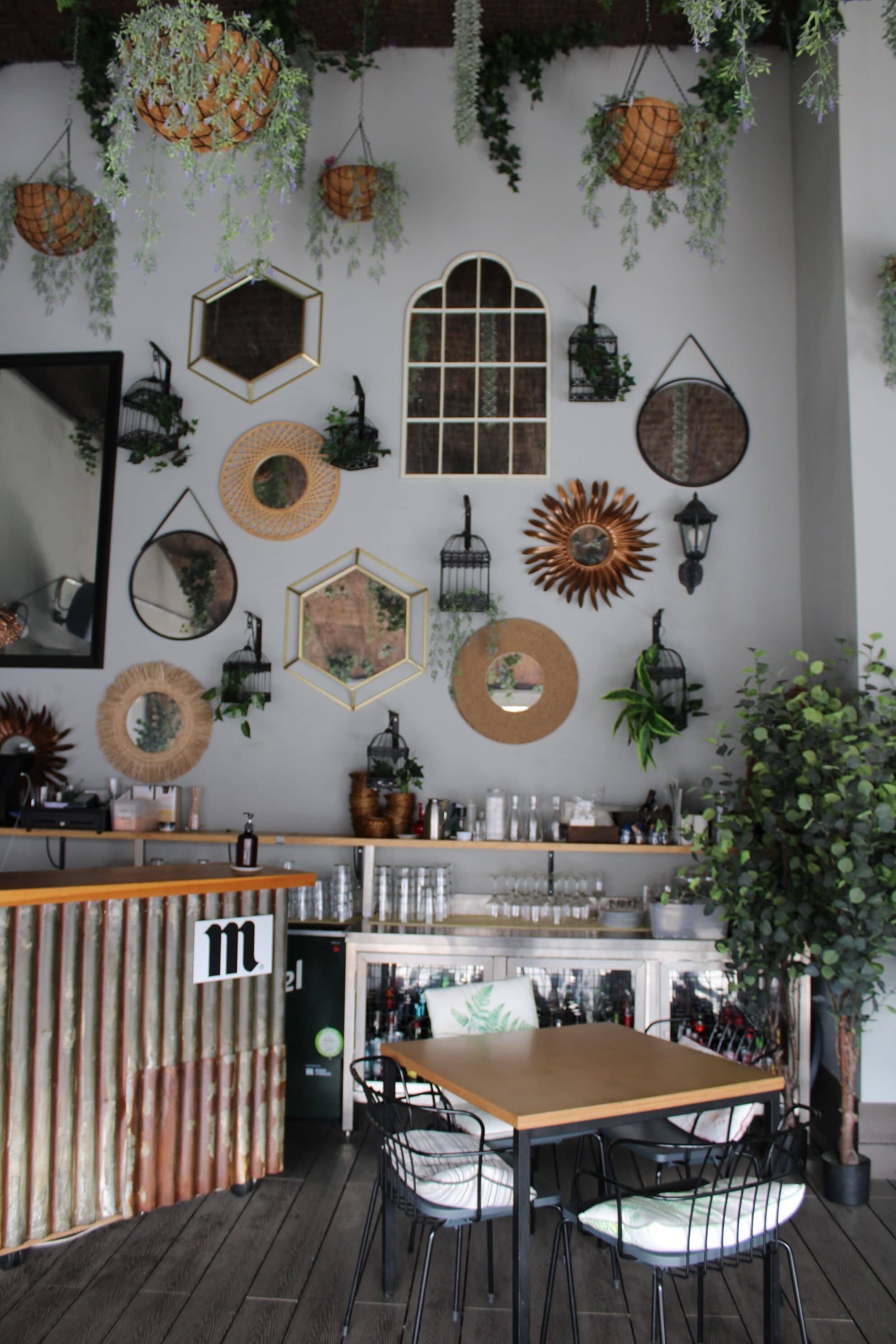 A cozy bar area with a wooden counter, multiple hanging planters, and a variety of wall decorations including mirrors and lanterns.