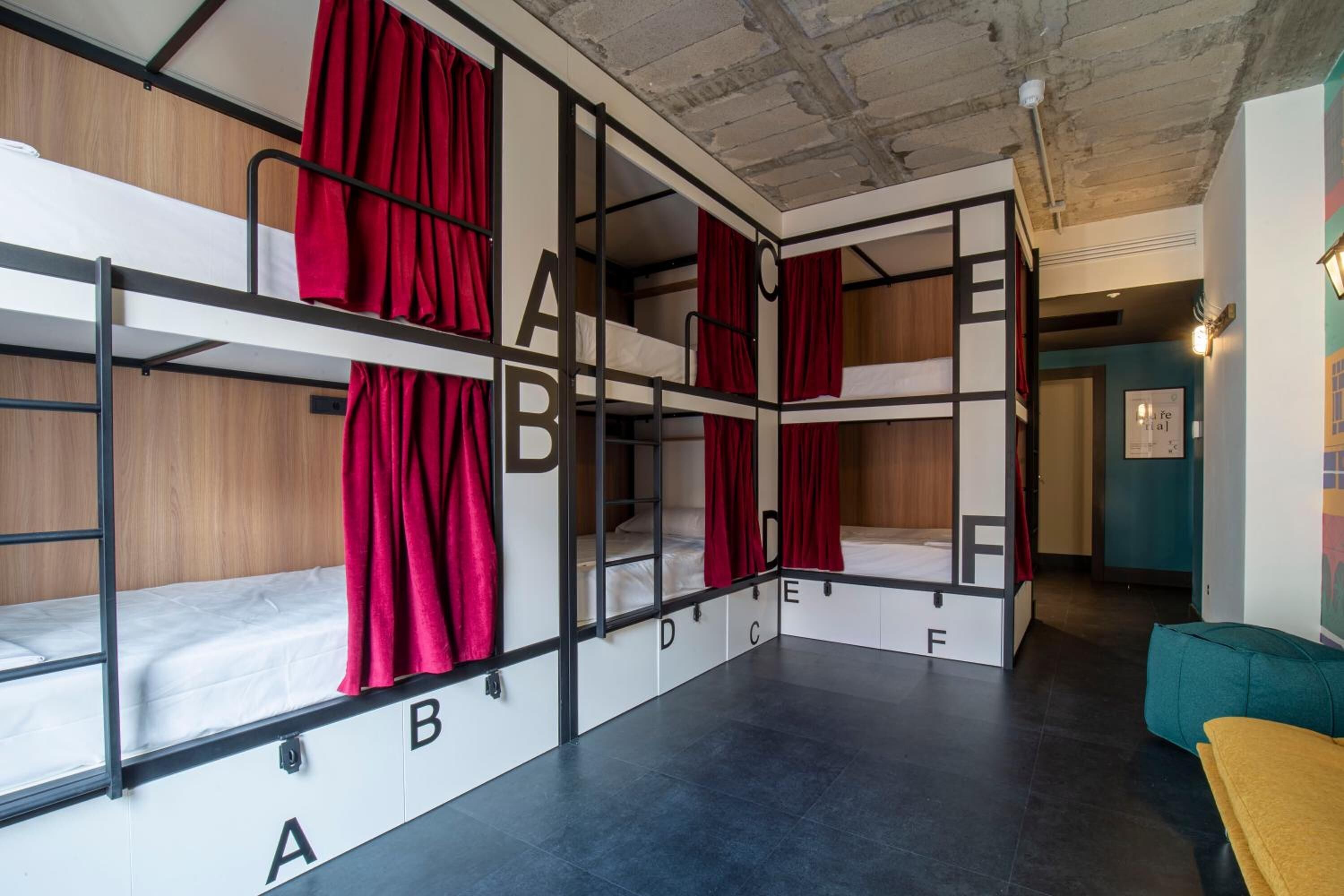 Habitación tipo dormitorio con literas etiquetadas de la A a la F, paredes de madera y suelo de baldosas.