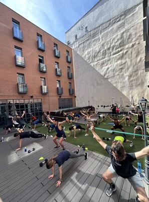 Een buitenfitnessles op een houten terras, met een bakstenen gebouw en een grote witte muur op de achtergrond.