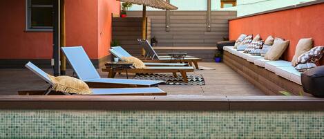 A sunlit outdoor lounge area with lounge chairs and a sofa, a small table, and a thatched umbrella.
