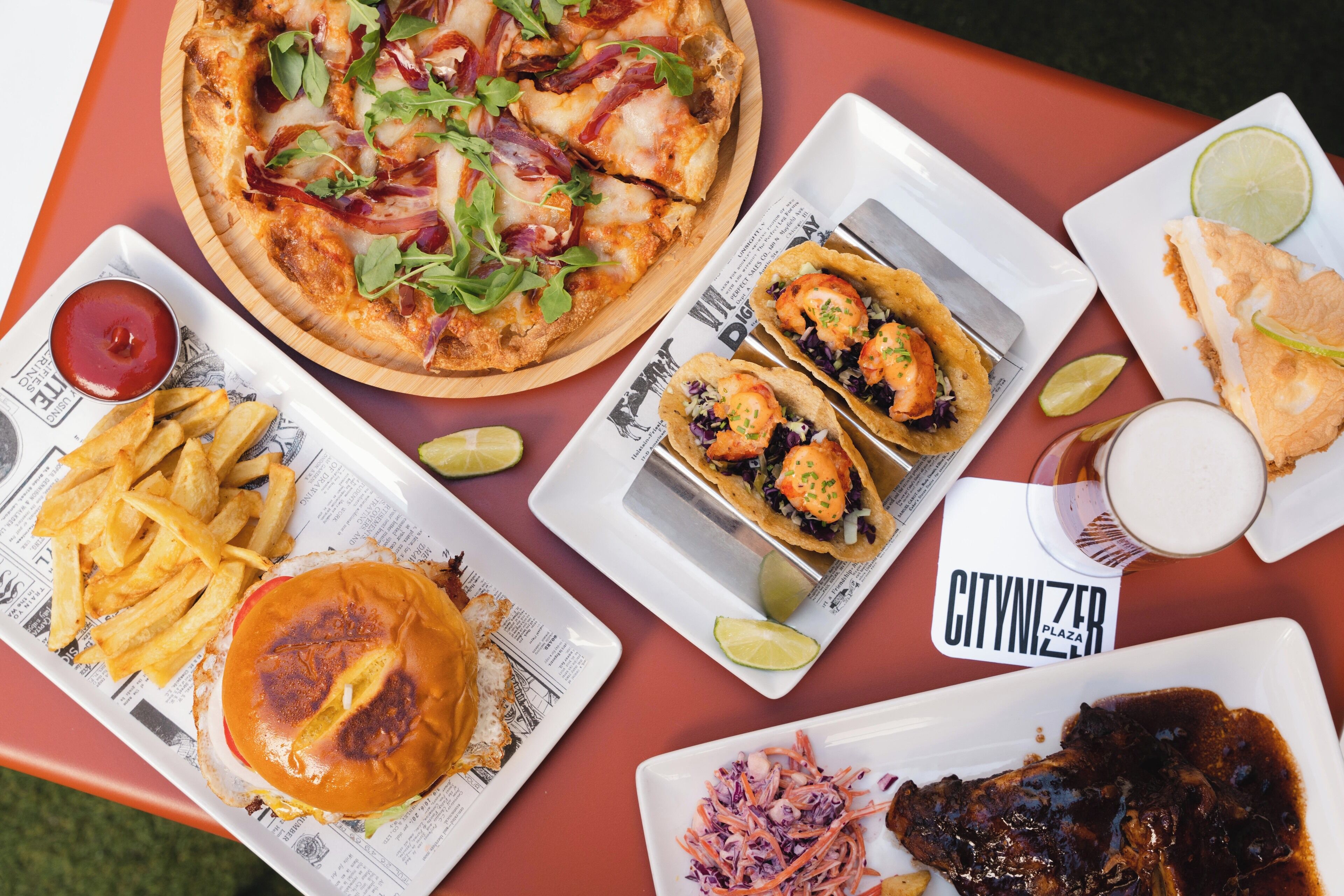 A table with various food items including a pizza, tacos, a burger, fries, coleslaw, and a steak.