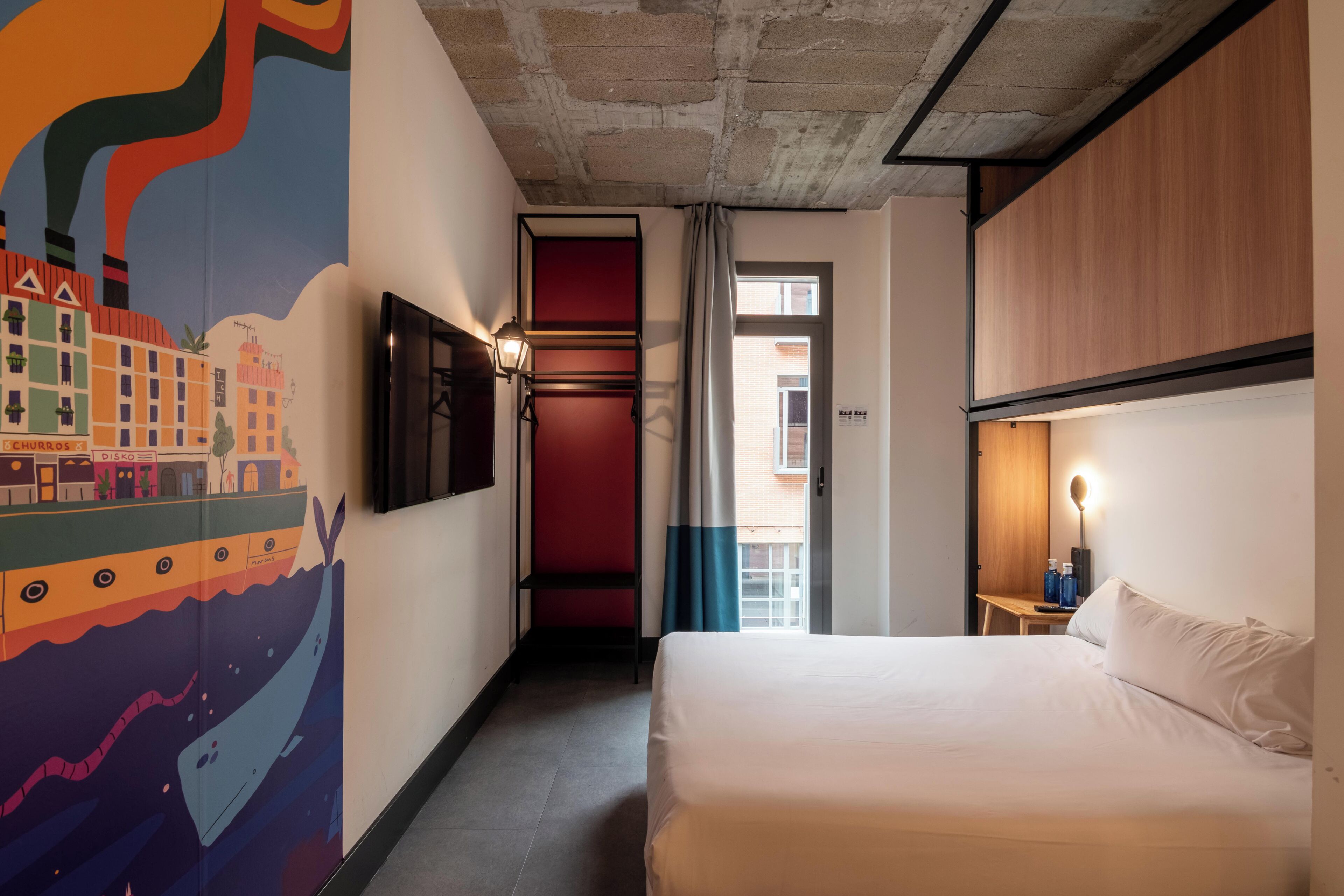 Una habitación de hotel con cama, televisión, un mural en la pared, ventana y puerta.
