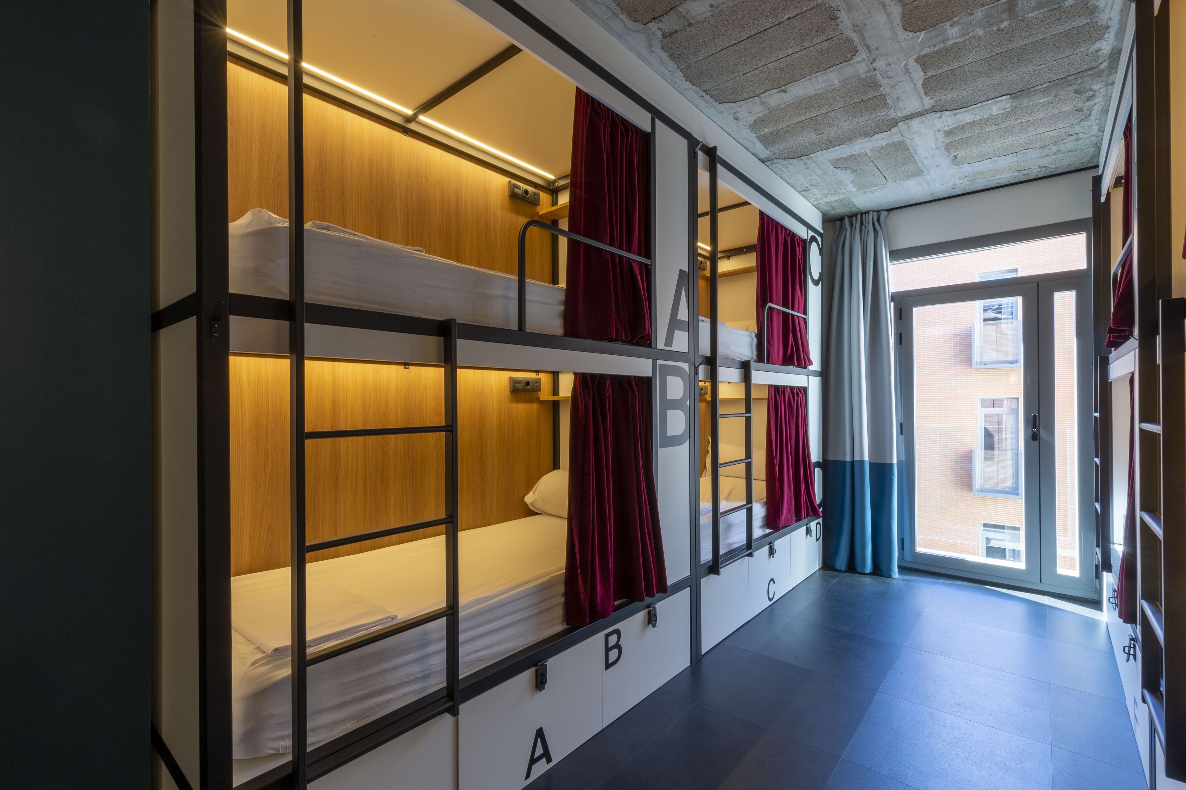 Ein Schlafsaalzimmer mit Etagenbetten, die mit A, B und C gekennzeichnet sind, Holzwandverkleidung und Blick durch ein großes Fenster.