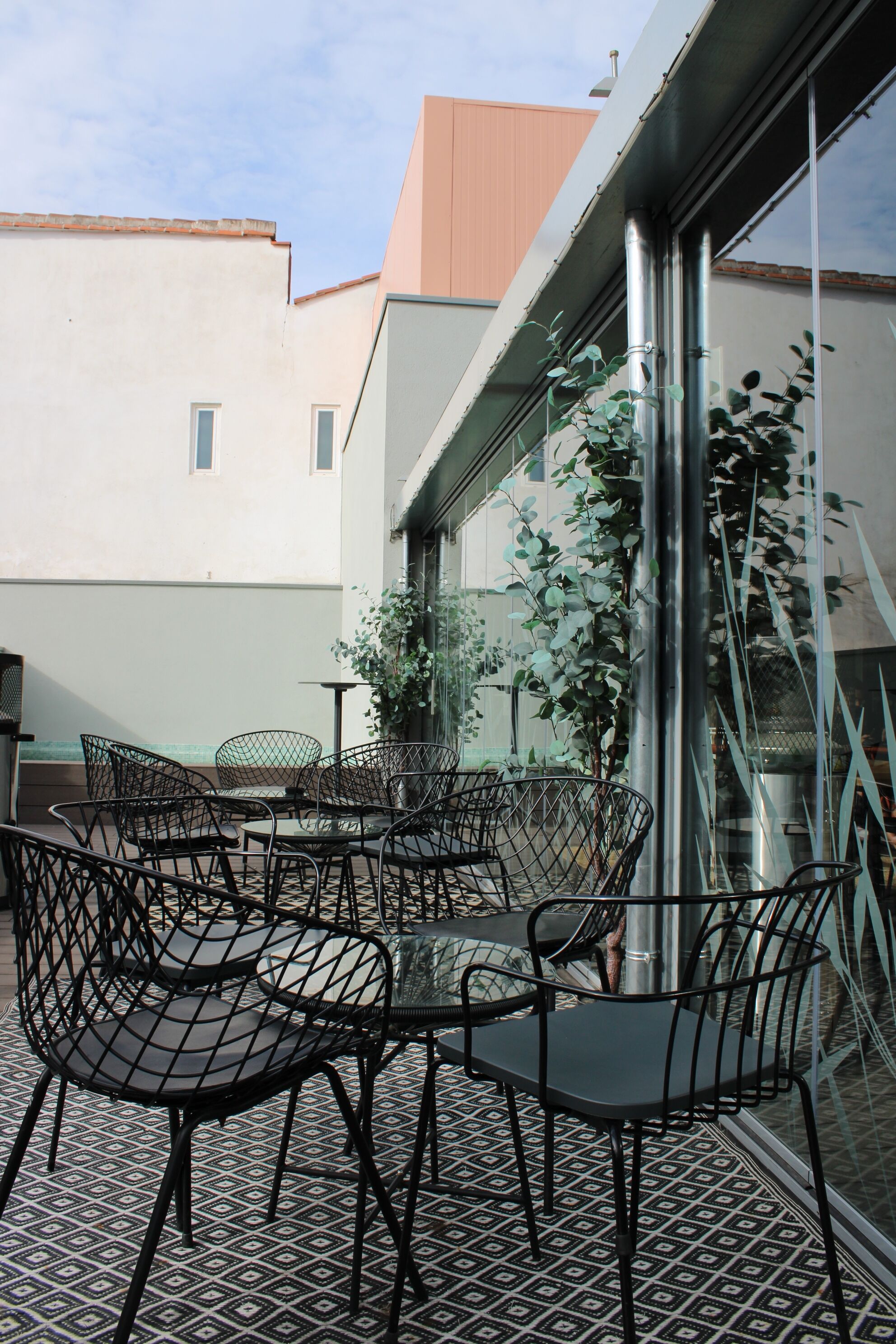 A terrace with metal chairs and tables, a glass railing, and a view of buildings in the background.