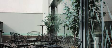A terrace with metal chairs and tables, a glass railing, and a view of buildings in the background.
