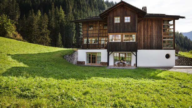 A wooden cabin with a balcony, large windows, and a stone wall foundation, surrounded by a grassy area and forested mountains.