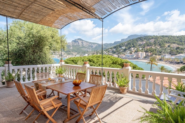 Une terrasse extérieure avec une table et des chaises, entourée de plantes en pot et offrant une vue panoramique sur une ville côtière et les montagnes.