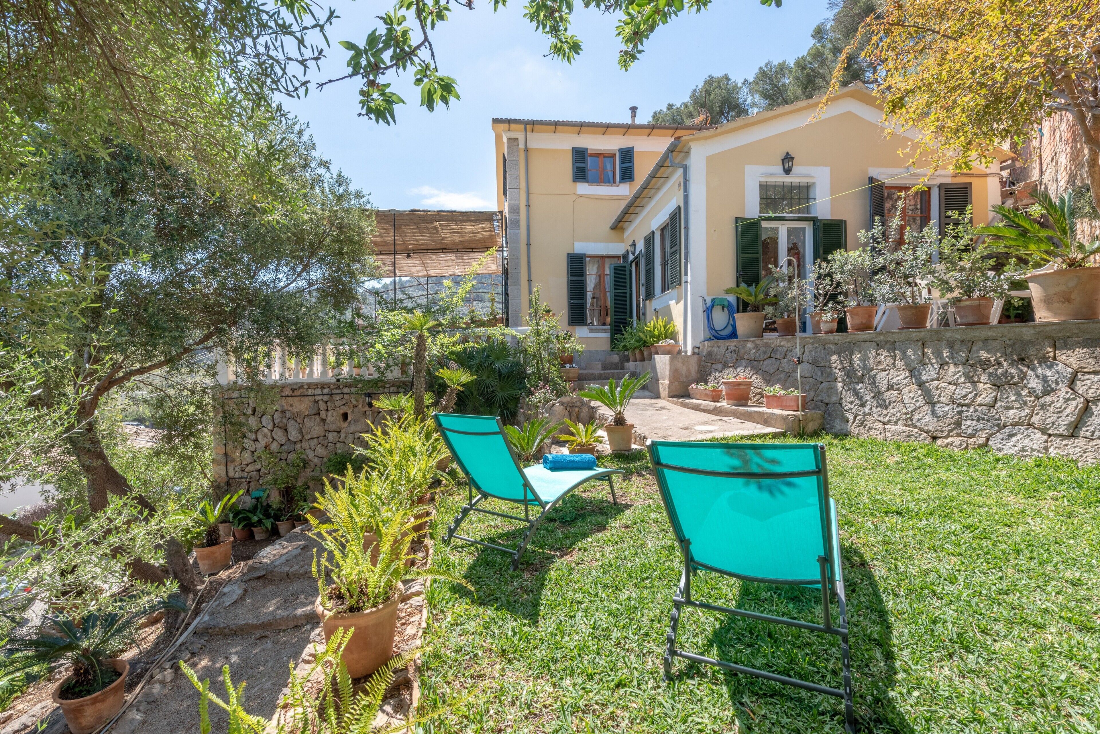 Una casa con un muro in pietra, un piccolo giardino con piante in vaso e due sedie verdi.