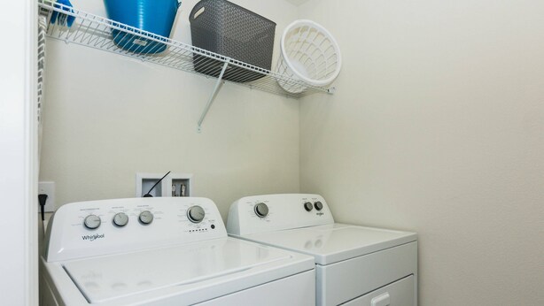 Un espace buanderie équipé de deux lave-linge et deux sèche-linge Whirlpool, d’une étagère en métal où sont rangés des paniers à linge et d’un mur blanc.