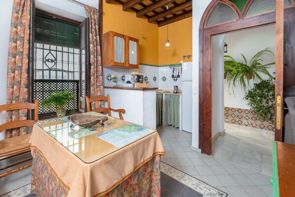 A dining area with a table set for two, a kitchen in the background, and a doorway leading to a balcony with plants.