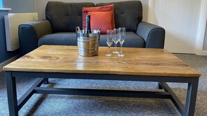 A wooden coffee table with a metal bucket containing a bottle and glasses, placed in front of a grey sofa with a red pillow. - The Roade House Rooms (Northampton)
